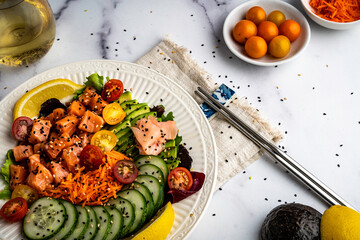 Salmon poke bowl with mixed salad greens, avocado, cucumber, carrots, and grape tomatoes. Garnished with black and white sesame seeds.