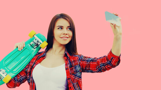 Portrait of young woman taking selfie with smartphone holding skateboard on pink background