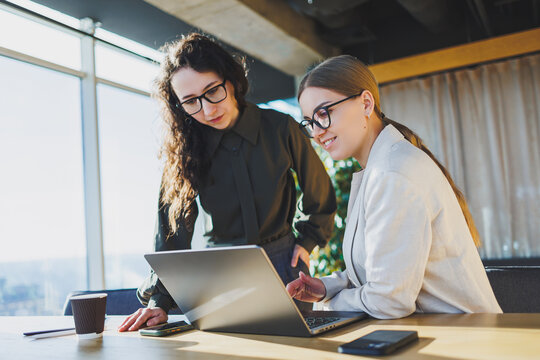 Smiling Female Colleagues Working On New Project At Laptop, Working In Modern Spacious Office With Large Windows. Woman In Glasses And Casual Clothes In The Office.