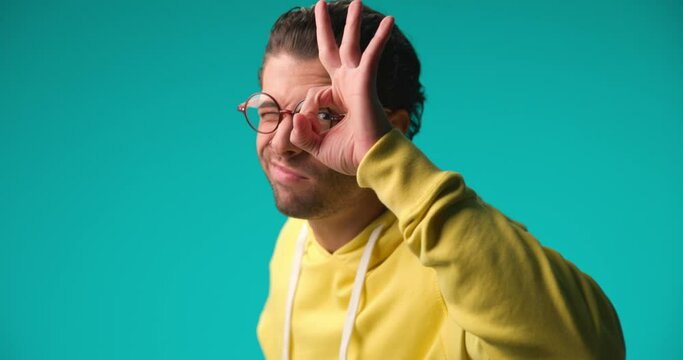 curly hair guy with glasses making ok gesture and looking through, being happy and smiling on blue background in studio