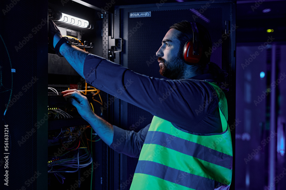 Wall mural Side view portrait of bearded man as network technician connecting cables and wires while repairing server in data center lit by neon light