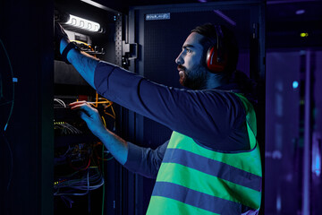 Side view portrait of bearded man as network technician connecting cables and wires while repairing server in data center lit by neon light