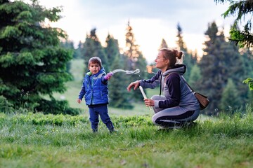 Mother plays with son on a lawn and teaches him to blow soap bubbles against the backdrop of forest