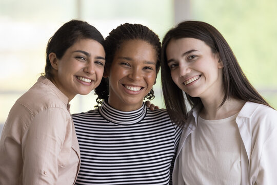 Happy diverse female business team posing indoors, hugging, standing close, looking at camera, with toothy smiles. Best friends women, business colleagues, successful teammates head shot portrait