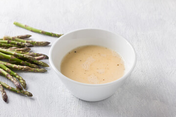 White ceramic bowl of beaten eggs with milk and fresh asparagus shoots on a light gray background. Cooking a delicious homemade omelet