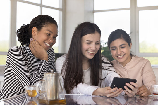 Happy multiethnic female friends talking on video call, using smartphone for communication, sitting together at table, watching online content on social media, smiling, laughing
