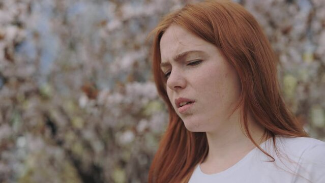 Close up of upset female having pollen allergy symptoms during spring outdoors. Frustrated caucasian lady rubbing her eyes while standing in front of blooming tree.