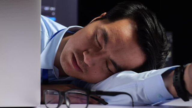 Close Up Of Asian Male Programmer Sleeping While Writing Code By A Laptop Using Multiple Monitors Showing Database On Terminal Window Desktops In The Office
