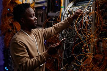 Side view portrait of black young man as system administrator fixing server with messy cables and...