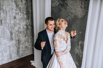 Stylish young groom in a blue suit and a beautiful blonde bride in a white lace dress are dancing,...