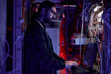 Side view portrait of bearded system administrator setting up server network and using laptop in neon light