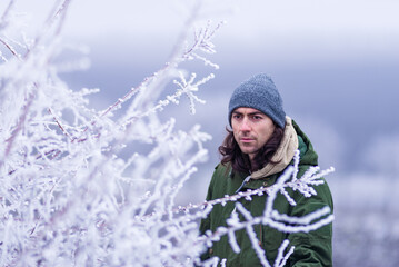 Fototapeta na wymiar close up frost man male young 30 years old long hair snow a winter day forest branches hoarfrost concept green coat park cold snow nature gray hat outdoors snowy Moldova.