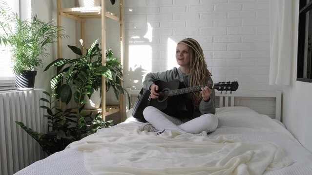 woman with dreadlocks on head singing song and playing acoustic guitar in sunny cottage bedroom, simple living and hobby relaxing