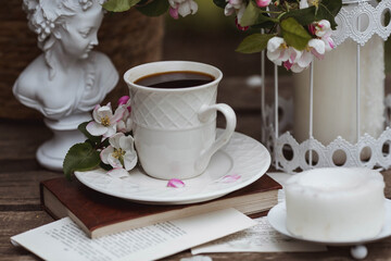 Obraz na płótnie Canvas A beautiful postcard. A white coffee cup with a saucer, a statuette, candles, a book and a vase with a bouquet of blooming apple trees. Beautiful still life. Spring time. The concept of 