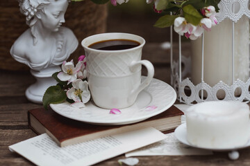 A beautiful postcard. A white coffee cup with a saucer, a statuette, candles, a book and a vase with a bouquet of blooming apple trees. Beautiful still life. Spring time. The concept of 