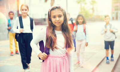 Portrait of positive schoolgirl standing near school, children on background