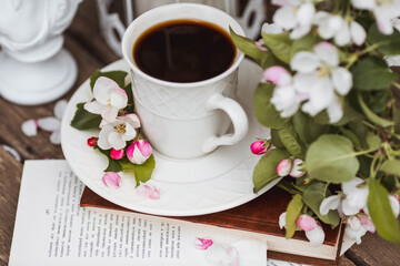 A beautiful postcard. A white coffee cup with a saucer, a statuette, candles, a book and a vase with a bouquet of blooming apple trees. Beautiful still life. Spring time. The concept of 