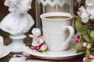 A beautiful postcard. A white coffee cup with a saucer, a statuette, candles, a book and a vase with a bouquet of blooming apple trees. Beautiful still life. Spring time. The concept of "Good morning"