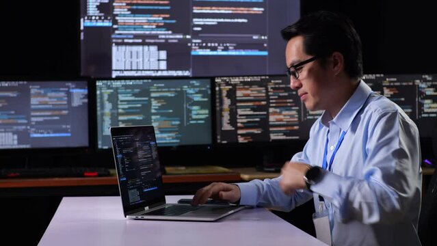 Side View Of Asian Male Programmer Walking Into The Working Space To Write Code By A Laptop Using Multiple Monitors Showing Database On Terminal Window Desktops In The Office
