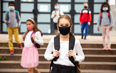Portrait of positive schoolgirl in mask standing near school, children on background