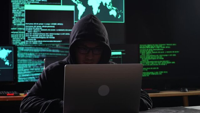 Asian Male Hacker In A Hood Works On A Laptop With Maps And Data On Display Screens Of Multiple Monitors In A Dark Office Room
