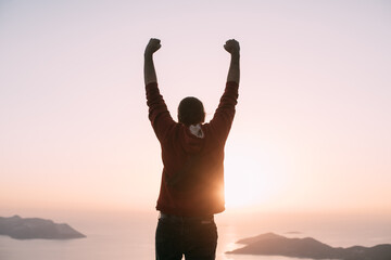 A young man meets the sunset, raising his hands in a victorious gesture on a mountain by the sea.