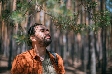 Portrait of a young man with a backpack on a walk in a pine forest on a sunny day. The guy breathes fresh air in a coniferous forest