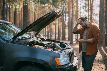 A frustrated man with a phone in his hands near a broken car with an open hood far outside the city...