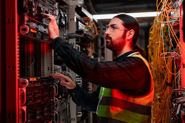 Side view portrait of bearded network technician inspecting servers and doing maintenance work in data center lit by red neon light