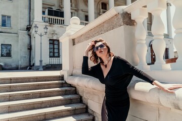 Fototapeta premium Stylish woman in the city. Fashion photo of a beautiful model in an elegant black dress posing against the backdrop of a building on a city street