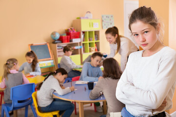 Upset girl standing in schoolroom on background with pupils studying with teacher