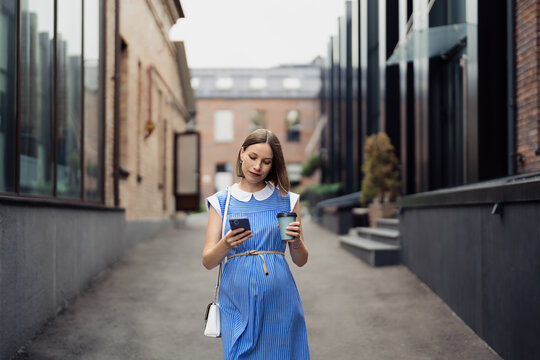 Busy modern pregnant woman walking among office buildings using a smartphone. Woman in a blue vintage dress and white purse using phone on the go