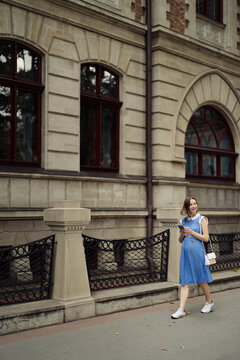 Beautiful young pregnant woman in a blue dress walking through the city streets using a smartphone.