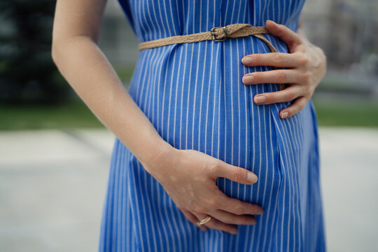Close up pregnant woman in a blue dress touching her tummy while resting in the city. Hands touch belly