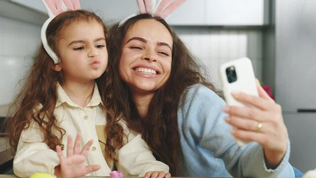 Happy mother and daughter doing selfie with easter eggs, wearing bunny ears, have beauty and fun day together at home. Concept of childhood, technology, family weekend