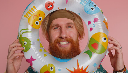 Close-up oortrait of funny young bearded hippie redhead tourist man looking out of rubber swimming ring hole smiling enjoying summer vacation time. Travel, recreation, sea journey. Pink background