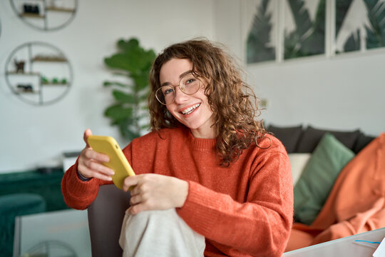 Smiling Young Pretty Woman Sitting At Home Using Apps On Cell Phone Technology, Happy Lady Holding Smartphone In Hands, Looking At Camera, Relaxing With Cellphone In Hands Checking Cellular Device.