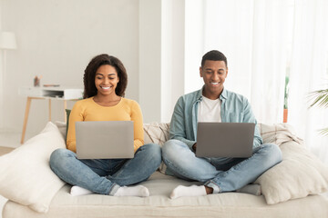 Cheerful young african american guy and lady with laptops work, surfing in internet, sit on sofa