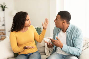 Angry sad emotional young african american lady and male screaming, gesturing, sitting on sofa