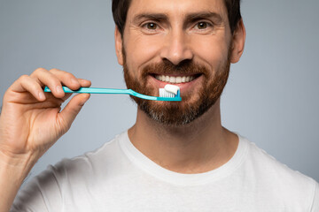 Happy caucasian man cleaning teeth with toothbrush and smiling, standing over grey studio background
