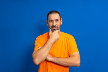Curious handsome bearded latino man thinking with hand touching chin on isolated studio blue background.