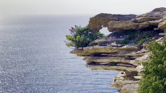 Elegant hole in a prominent strata layered piece of stone protruding from the rocky cliff of Accore coast near Bonifacio (Corsica, France), with green vegetation, horizon and sea waves in background