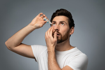 Handsone bearded caucasian man applying ophthalmology eyedropper, standing on grey studio background