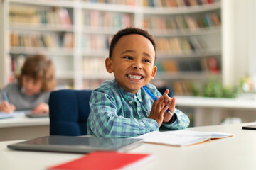 Happy african american primary school boy sitting at desk in classroom, writing in notebook and...