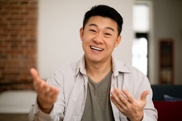 Hello. Portrait of happy mature asian man gesturing and talking at camera, sitting on sofa at home