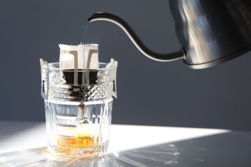 Pouring hot water into glass with drip coffee bag from kettle on light grey table, closeup