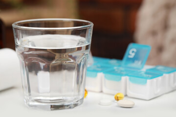 Plastic pill box with different medicaments indoors, focus on glass of water. Space for text