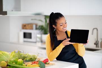 Happy young african american woman in apron show tablet with empty screen at table with fresh vegetables