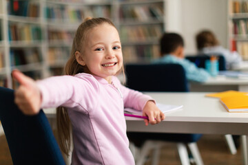Happy caucasian schoolgirl gesturing thumbs up and smiling to camera, sitting at school in classroom, free space