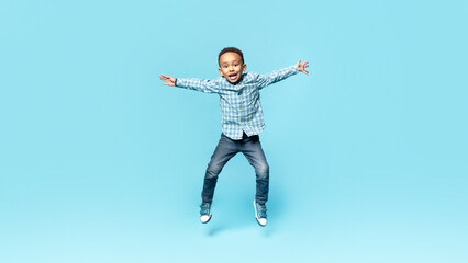 Fototapeta na wymiar Happy little african american boy posing in mid air, jumping and spreading arms over blue studio background, full length